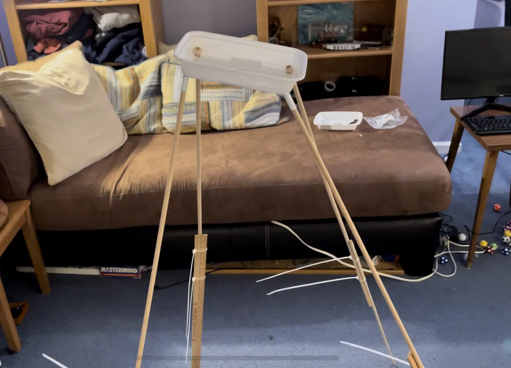 The rebuilt camera mount standing on the floor: taped dowel legs holding a plastic takeout container for the camera at the top